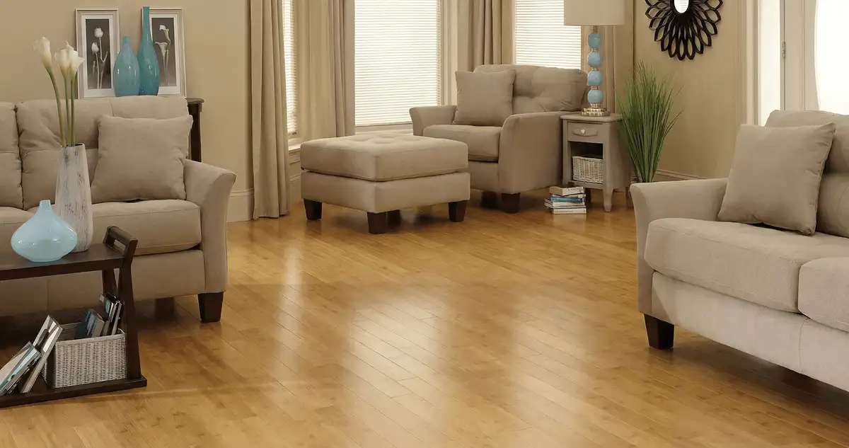 A living room with professionally installed bamboo flooring, beige sofas, a matching ottoman, a side table with a blue lamp, vases, framed pictures, and large windows with blinds and curtains in San Antonio, TX